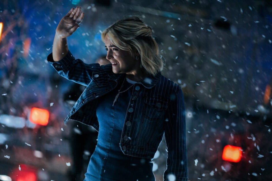 Person with blonde hair wearing a denim jacket and dress smiles while raising a hand, surrounded by falling snow.