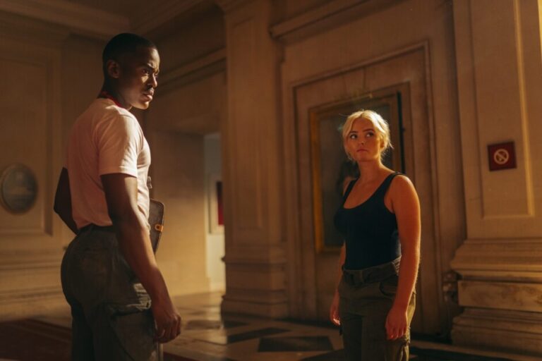 ‘Doctor Who: Empire of Death’ Season 1, Episode 8 Review – An Explosive, Emotionally Charged Finale Two individuals stand in a dimly lit interior with paneled walls and framed paintings, engaged in conversation. The person on the left wears a white t-shirt and dark pants, the person on the right wears a black top and grey pants.