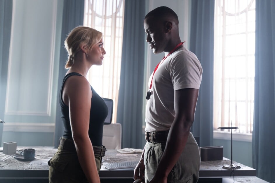 A woman and a man stand facing each other in a room with tall windows and blue curtains. The woman wears a black tank top and the man wears a white t-shirt with a red lanyard around his neck.