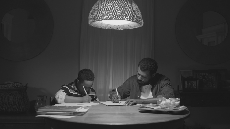A man and a boy sit at a table under a lamp, concentrating on writing or drawing on papers in a dimly lit room.