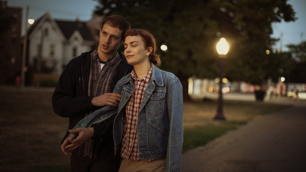 A man and woman walk on a park path at dusk. The man supports the woman, whose eyes are closed. She wears a denim jacket, and they both appear relaxed. Streetlights and trees are visible in the background.