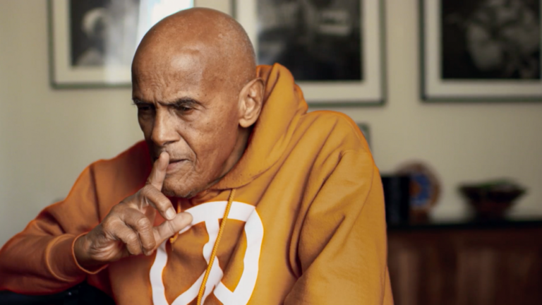 An elderly man with a shaved head wearing an orange hoodie with a peace symbol holds a finger to his lips, sitting in an indoor setting with framed pictures on the wall behind him.