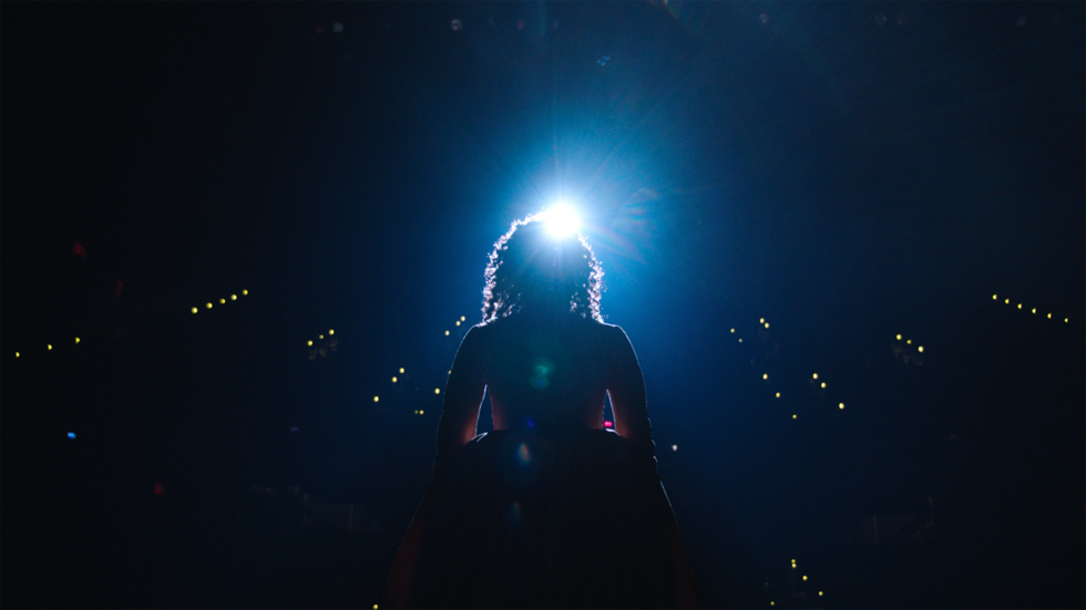 A person with curly hair stands on a stage, illuminated from behind by a bright light, facing an audience in a dark auditorium.