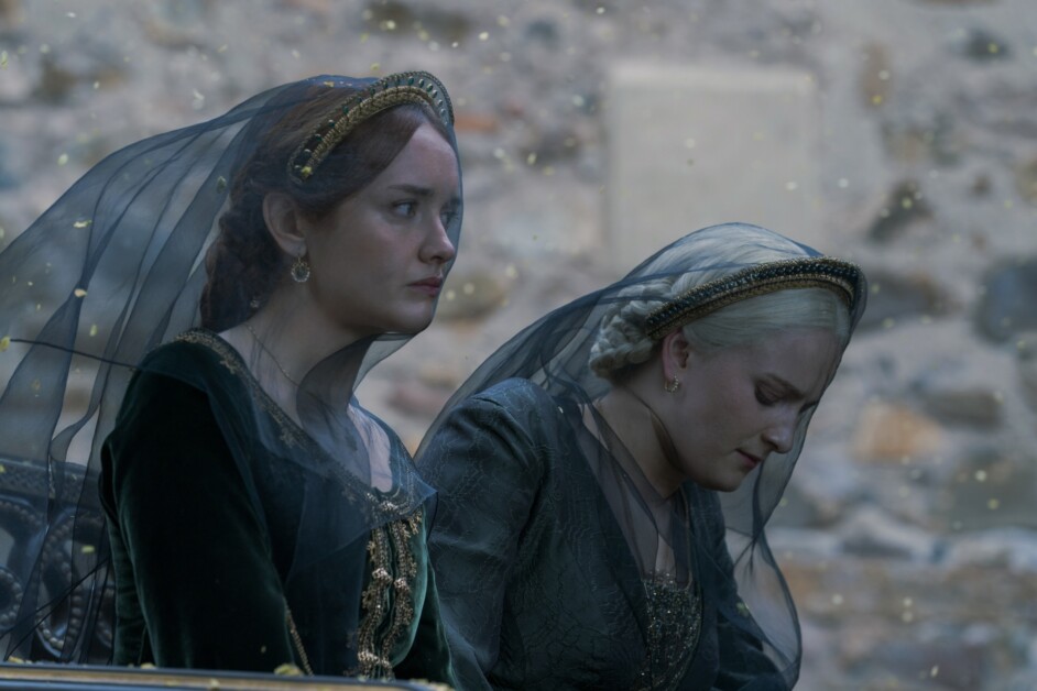 Two women wearing dark green medieval-style dresses and black veils sit with serious expressions against a stone wall background.