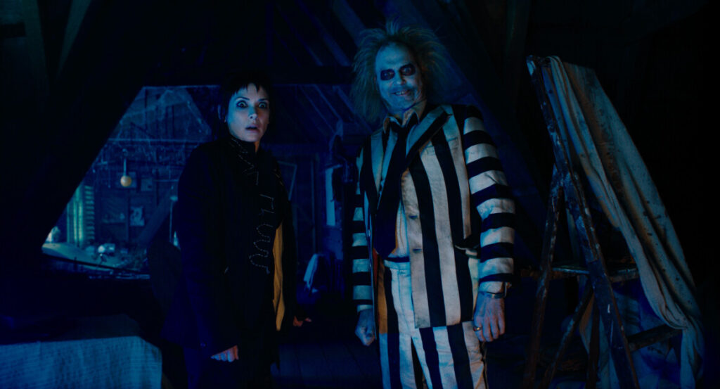 Two people in a dimly lit room, one in a black outfit with short dark hair, and the other dressed in a black-and-white striped suit, with pale skin and disheveled hair, standing side by side.