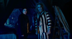 Two people in a dimly lit room, one in a black outfit with short dark hair, and the other dressed in a black-and-white striped suit, with pale skin and disheveled hair, standing side by side.