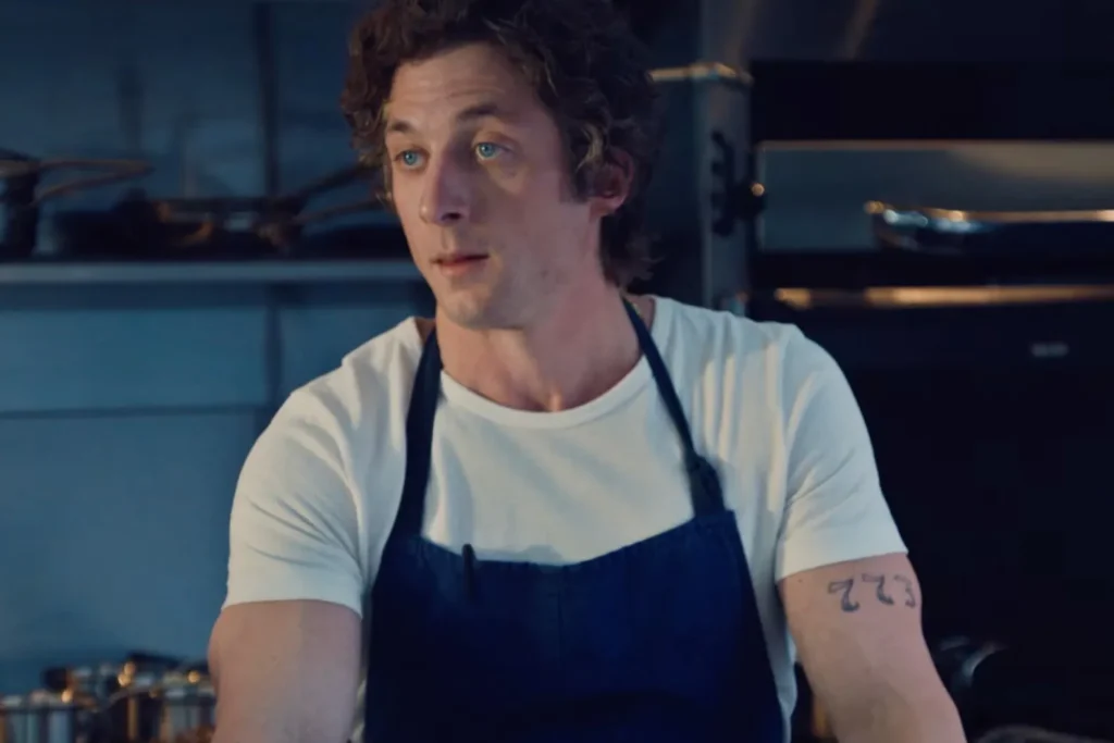 A person with curly hair wearing a white t-shirt and a dark apron stands in a kitchen, with cooking equipment visible in the background.
