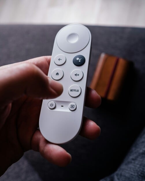 A hand holding a white remote control with buttons for power, volume, Google Assistant, and Netflix. A wooden object is blurred in the background.