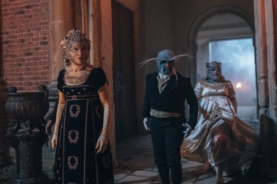 People in elaborate fantasy costumes walk through a dimly lit stone corridor, showcasing ornate wigs, makeup, and period-style clothing.