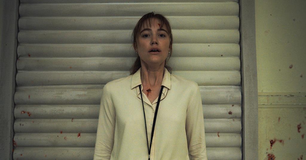 A woman with a lanyard stands against a wall with blood splatters, looking ahead with a serious expression.