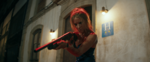 A person with disheveled hair and a determined expression holds a red shotgun under dim lights in front of a building with an "H" sign.