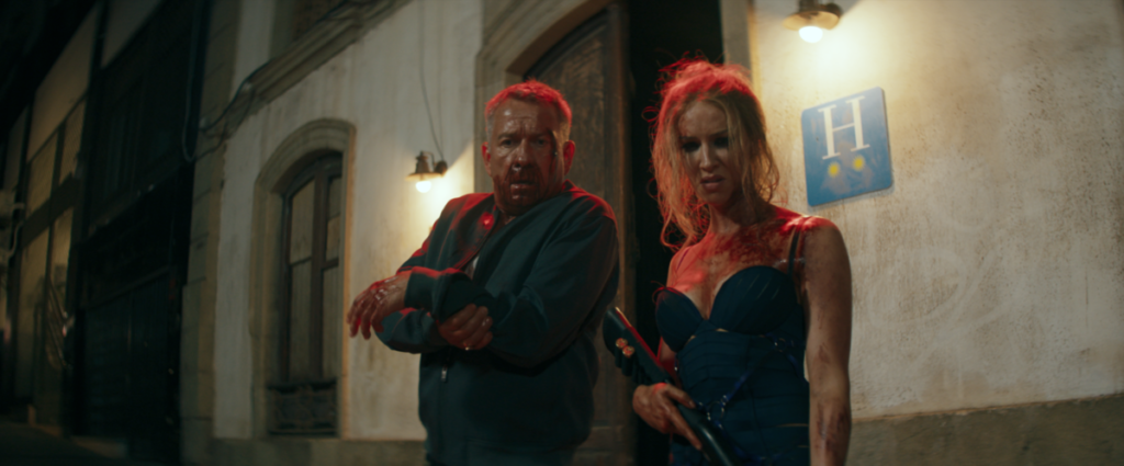 A man and a woman, both with bloodied faces, stand in a dimly lit street. The woman, holding a rifle, is dressed in a blue dress, while the man appears injured, holding his arm in pain.