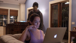 A person in a mask stands menacingly behind a woman who is focused on her laptop in a residential setting.