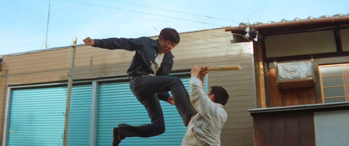 Two people engaged in a fight; one is airborne with a raised fist while the other, holding a wooden stick, defends. The scene is set outdoors near blue garage doors and wooden buildings.