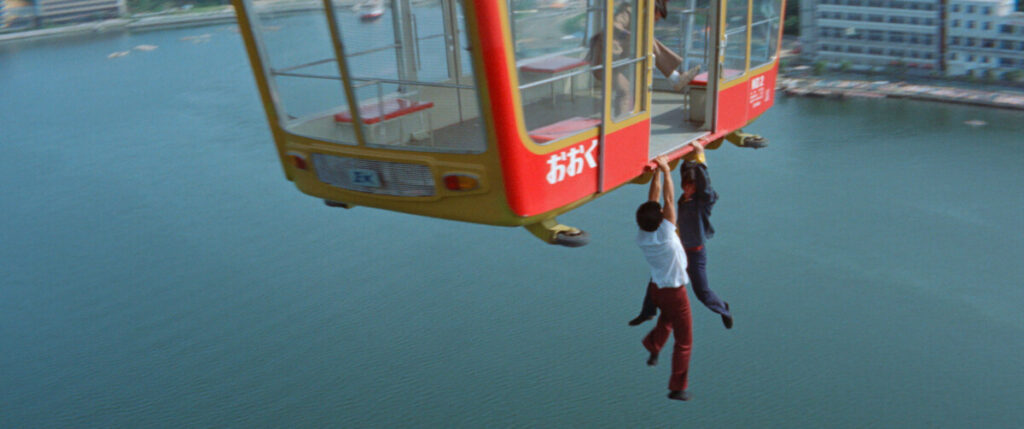 Two people hang from the underside of a red cable car suspended over a body of water with buildings in the background.
