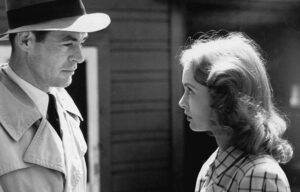 ‘Act Of Violence’ Blu-Ray Review – Sins Of The Past Refuse To Stay Buried In Gripping Thriller A man in a trench coat and hat looks intently at a woman with long hair who is wearing a checkered shirt, in a black and white photo.