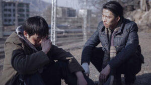 Two men sit outside on a rocky ground. One man, distressed, covers his face with his hand. The other, slightly dirty, faces him and appears to be speaking or comforting him. Urban buildings in the background.
