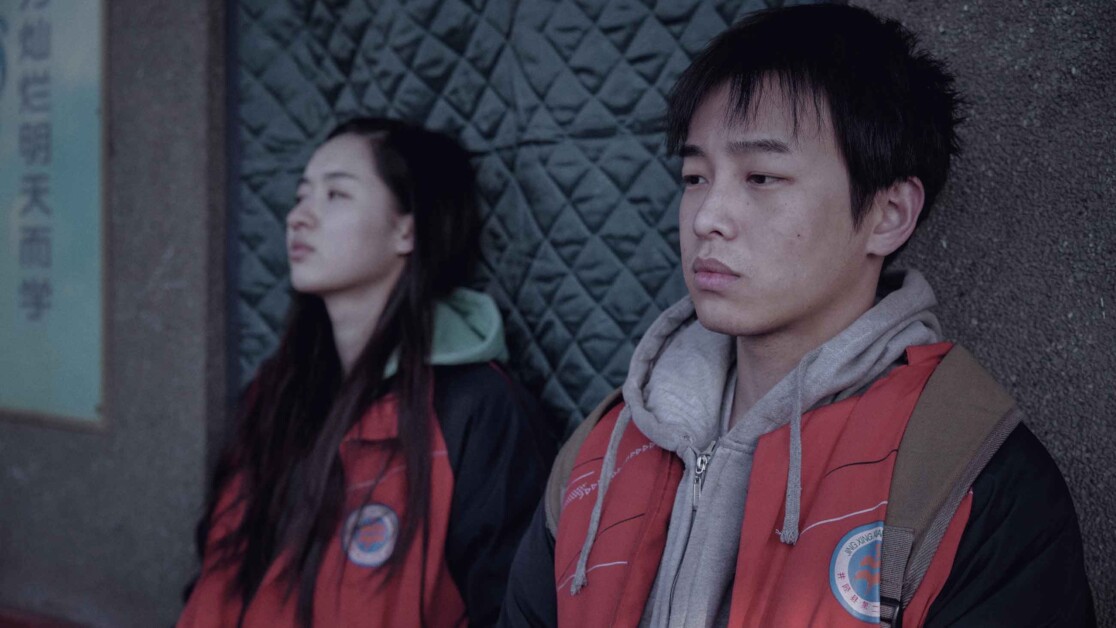 Two individuals wearing hooded jackets stand against a textured wall with a sign displaying some text in the background. Both appear to be looking off into the distance with neutral expressions.