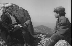 Two men, one seated and one kneeling, in conversation on a rocky cliff with distant mountains visible in the background. Both are dressed in heavy clothing and caps.