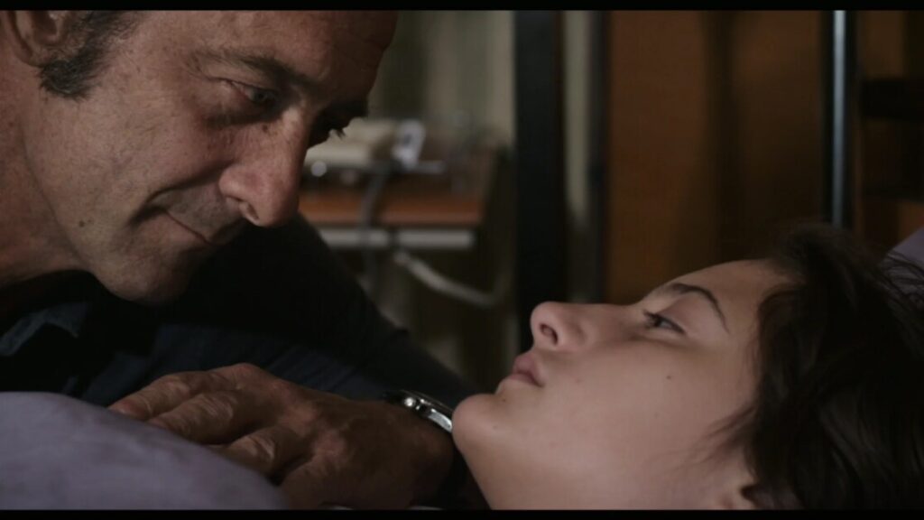 A man and a young person lie on a bed, looking intently at each other. A telephone and some cables are visible in the background.