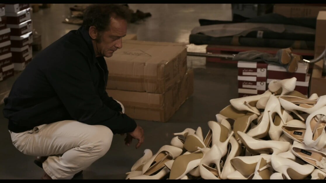 A man wearing a dark jacket and light pants crouches next to a pile of white shoes in a storage room with boxes.