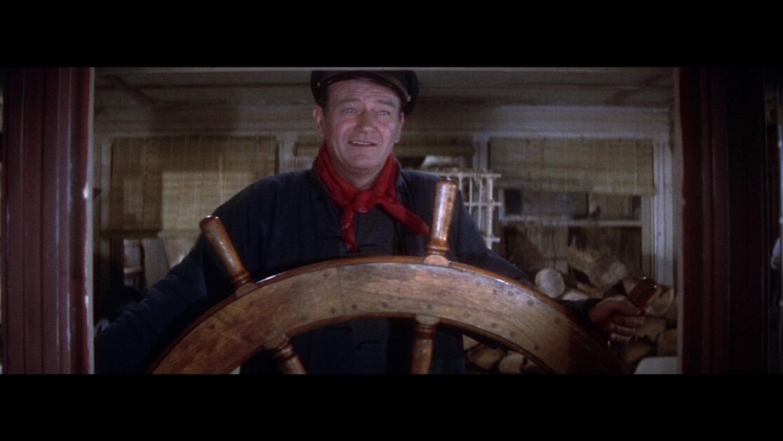 A man wearing a hat and red scarf stands behind a wooden ship's wheel indoors.