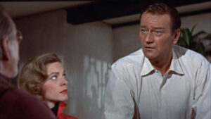 ‘Blood Alley’ Blu-Ray Review – John Wayne Leads Suspenseful Rescue Tale A man and a woman in a room, both looking in the same direction, with the man appearing to be speaking.