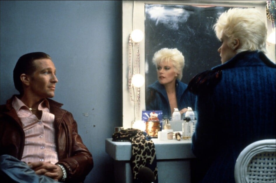 A person sits and looks at another person with blonde hair who is gazing at their reflection in a mirror on a vanity table with various items.