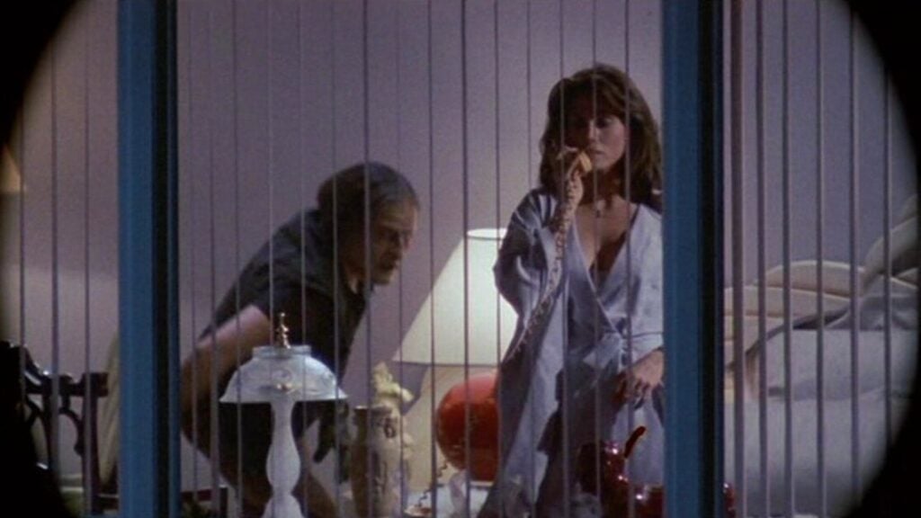 A man and woman are visible through window blinds. The woman is on the phone wearing a robe, while the man is bending forward beside her.