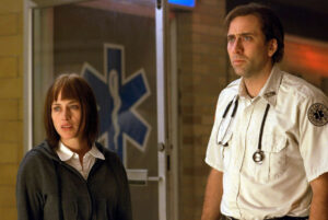 Two people, one wearing a grey hooded sweatshirt and the other in a paramedic uniform, standing indoors in front of a glass door with a medical cross symbol.