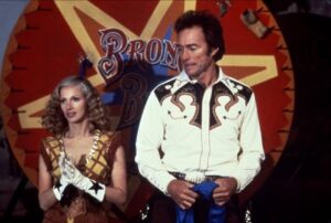 ‘Bronco Billy’ Blu-Ray Review – Clint Eastwood Crafts A Charming Tale Of Found Family A man and woman in western attire stand in front of a colorful "Bronco Billy" sign. The man wears a white shirt with black patterns; the woman has a yellow dress with gloves.