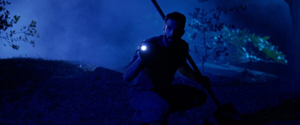A man holding a flashlight and a shovel kneels on the ground in a dark, misty, blue-lit forest.