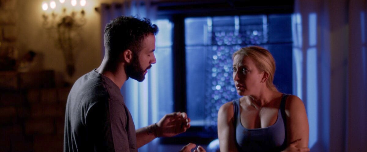 A man and woman engage in an intense conversation inside a dimly-lit room with a chandelier and blue light in the background.