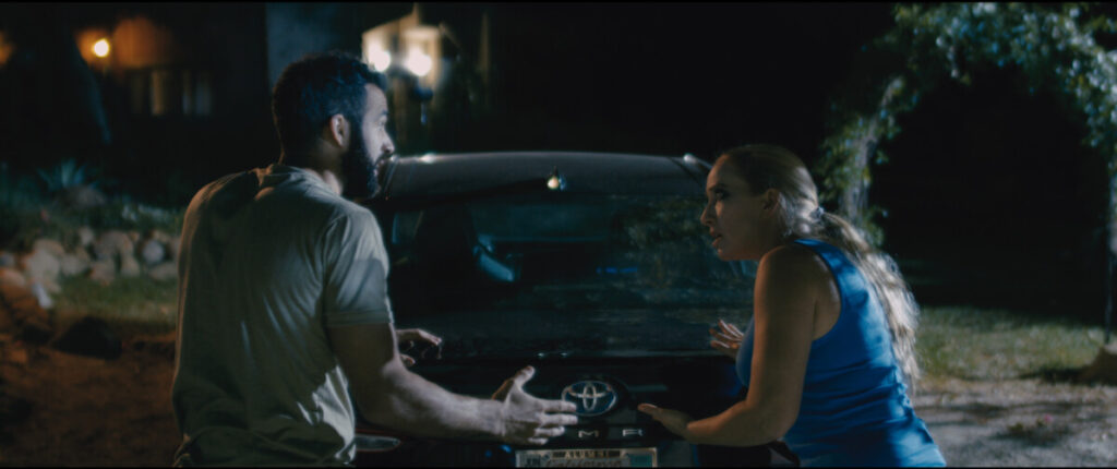 Two individuals, a man and a woman, are having an intense conversation while standing on opposite sides of a black Toyota car at night.