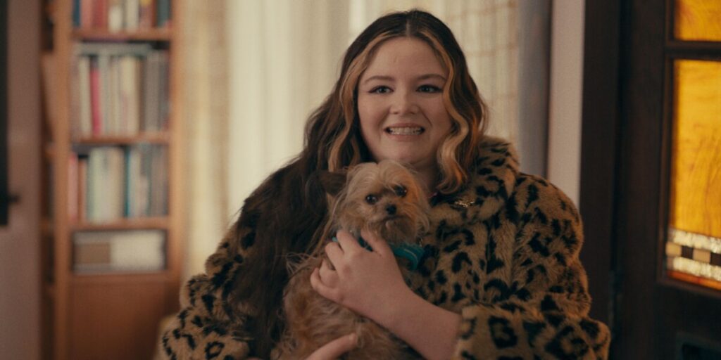 A woman wearing a leopard print jacket smiles while holding a small brown dog in her arms. A bookshelf and window are visible in the background.