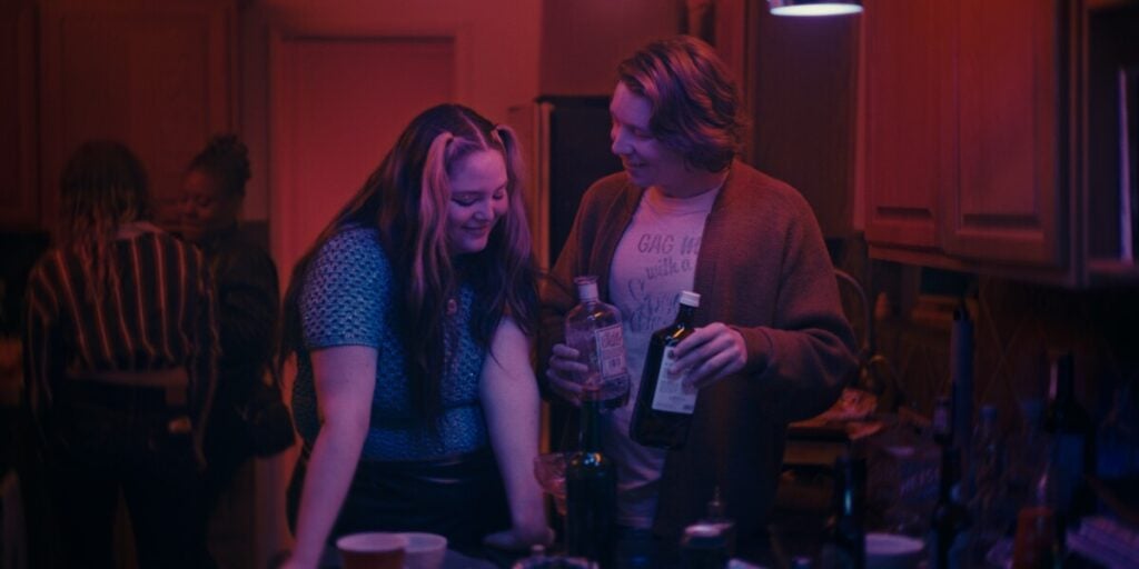 Two people stand close together in a dimly lit room, smiling. One person holds a liquor bottle and a cup, while the other looks down at the table. Others are in the background. The lighting is red and purple.