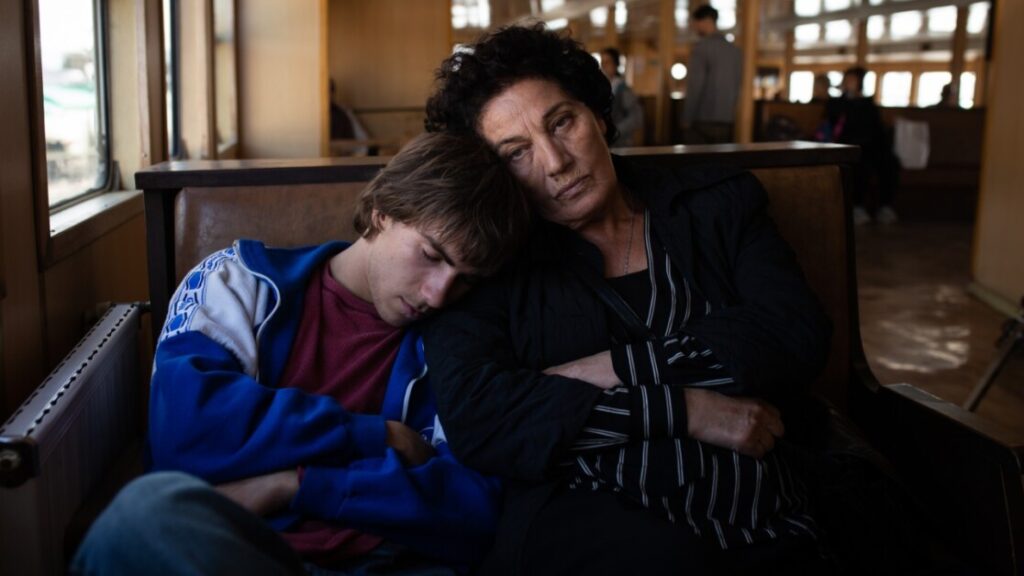 A young person in a blue jacket sleeps with their head resting on the shoulder of an older person wearing black, in a wooden seated area. The older person appears to be looking forward, with a neutral expression.
