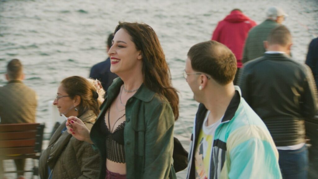 A group of people walking near a waterfront, with a woman in focus smiling and conversing with a man beside her. Other individuals are blurred in the background.