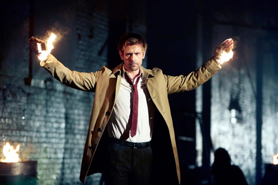 A man in a trench coat stands with arms outstretched, hands engulfed in flames, in a dimly lit, industrial setting.