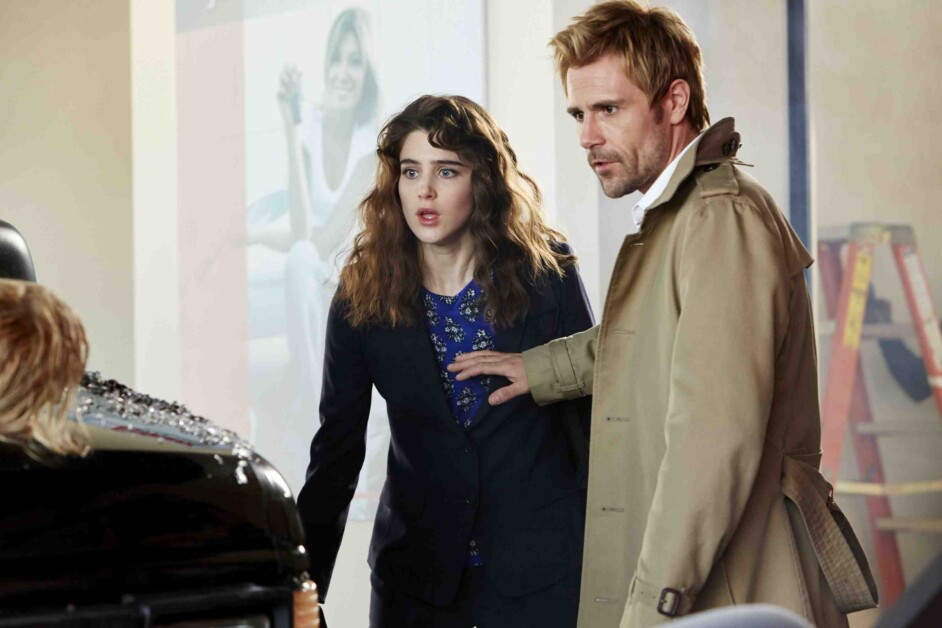 A man in a trench coat places his hand on a woman's shoulder. They both look concerned, standing next to a car with a shattered windshield. A person is visible inside the car.
