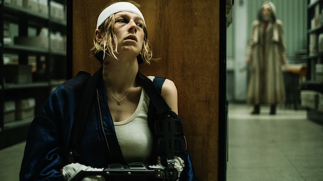 A person with a head bandage and arm cast, wearing a tank top, sits on the floor against a wooden wall in a storage room. In the background, another person in a long coat stands at a distance.