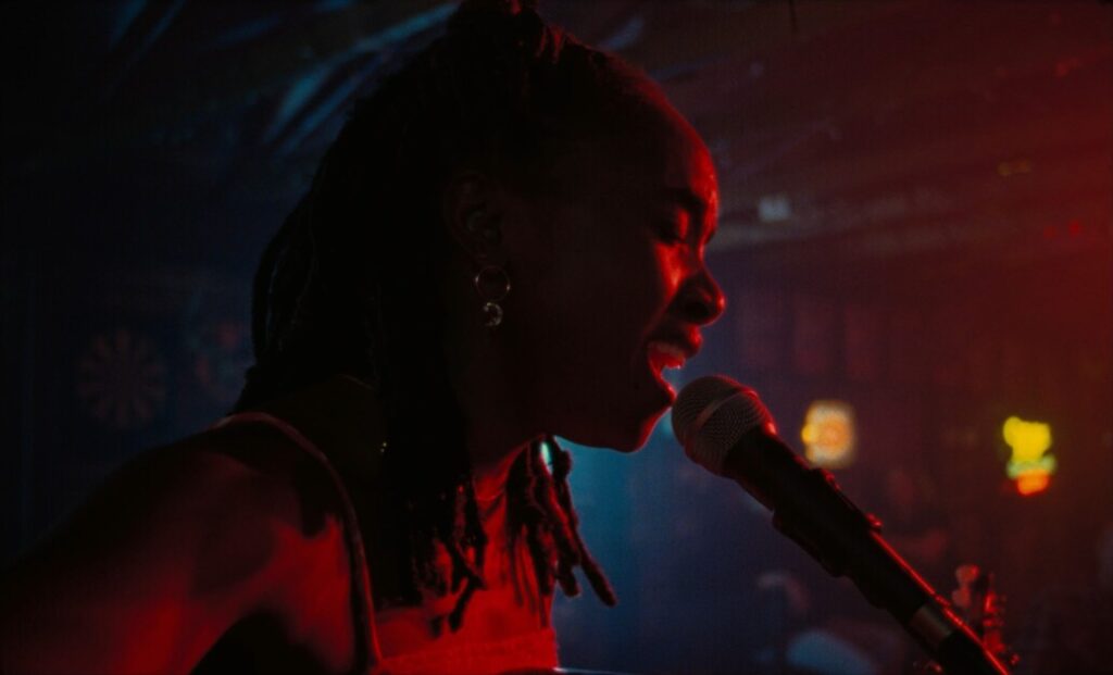 A person with long dreadlocks sings passionately into a microphone with red and blue lighting in a dimly lit venue.