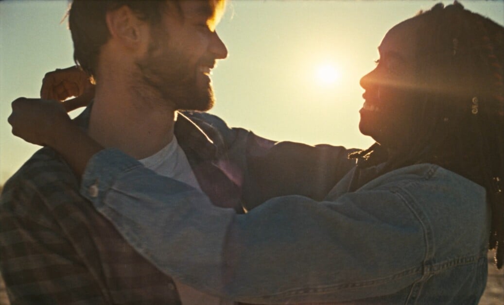 A man and woman, wearing casual clothes, embrace and look at each other with smiles. The sun is setting in the background, casting a warm light on the scene.