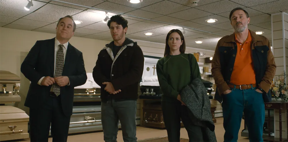 Four people stand in a showroom with caskets displayed behind them, appearing to listen intently. There is an older man in a suit, a younger man, a woman, and another man in casual attire.