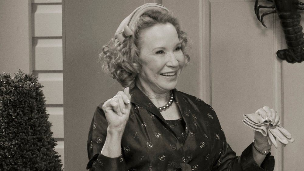 A woman with curly hair, wearing a dress and headband, smiles while holding gloves. She stands indoors near a topiary plant. The image is in black and white.