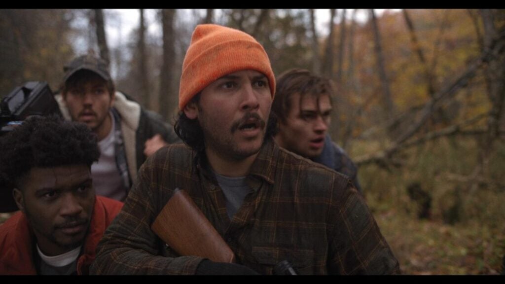 Four men in an outdoor wooded setting look ahead with concerned expressions. One man, wearing an orange beanie, holds a rifle.
