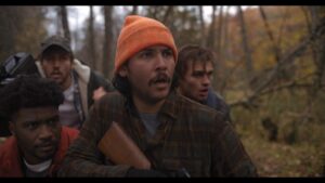 Four men in an outdoor wooded setting look ahead with concerned expressions. One man, wearing an orange beanie, holds a rifle.