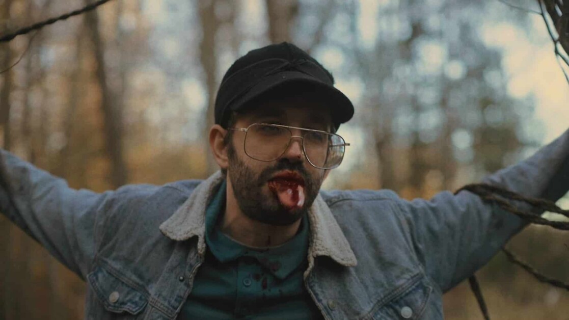 A man in a denim jacket and glasses, with blood on his mouth, stands in a forest with his arms outstretched.