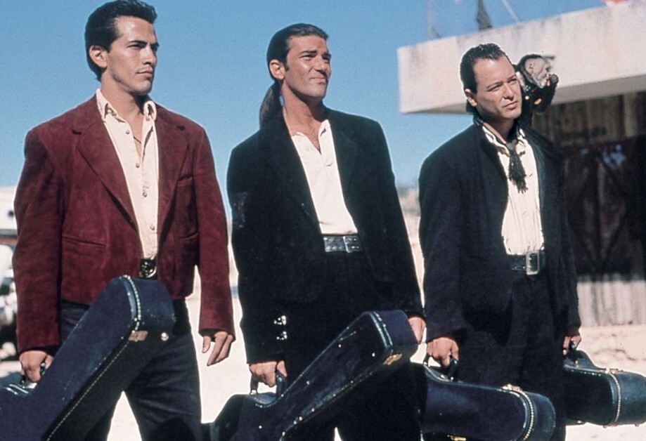 Three men stand side by side outdoors, each holding a guitar case. The man on the left wears a red jacket, the man in the middle wears a black suit, and the man on the right wears a dark blue jacket.
