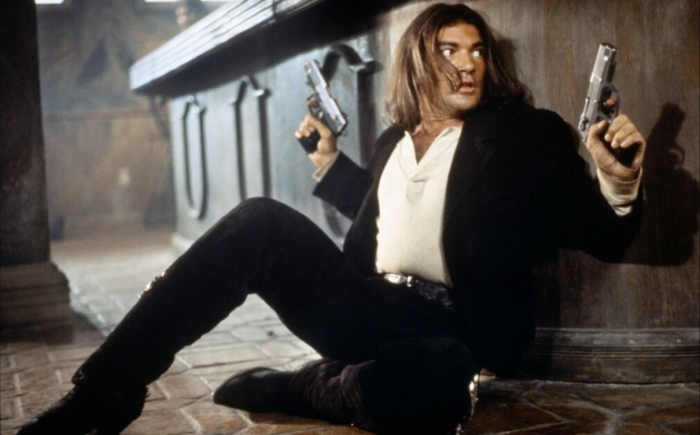 A man with long hair and a black outfit sits on the floor against a counter, holding two pistols and looking alert.